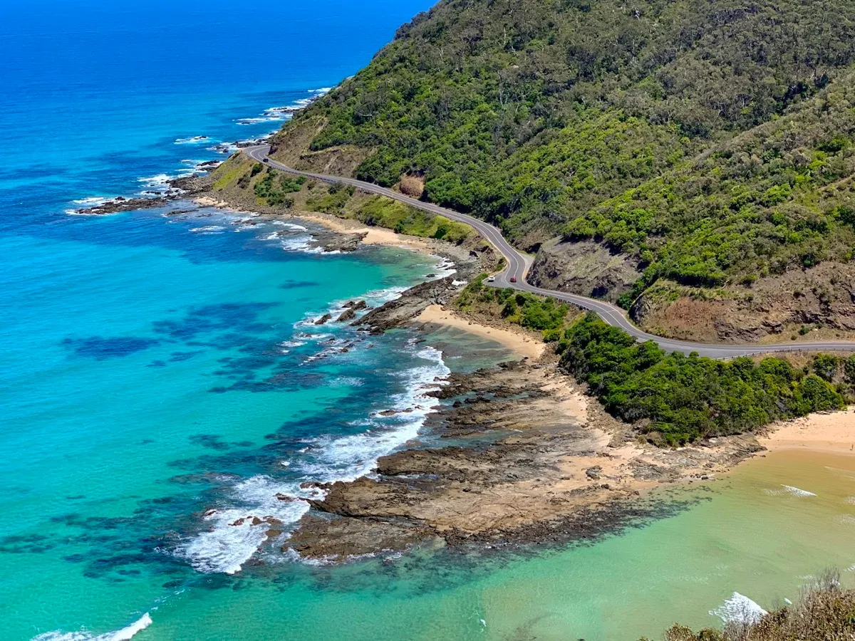 Aerial view of a coastal road winding between green hills and turquoise sea