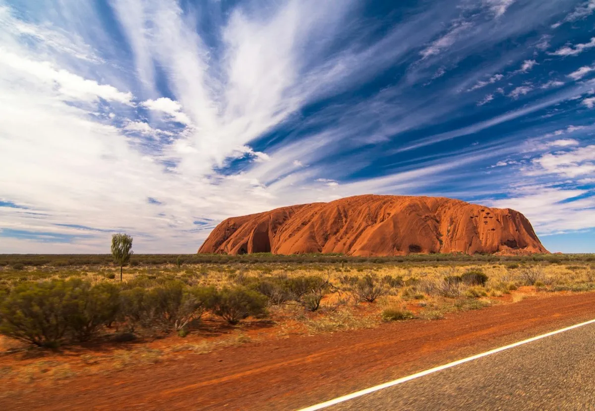 Uluru rising above red desert scrub beside an empty road